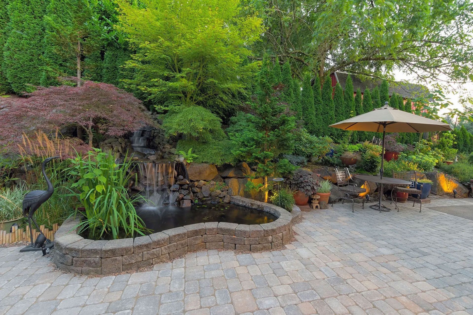 A landscaped backyard with a waterfall, pond, patio, table, and umbrella; lush greenery.