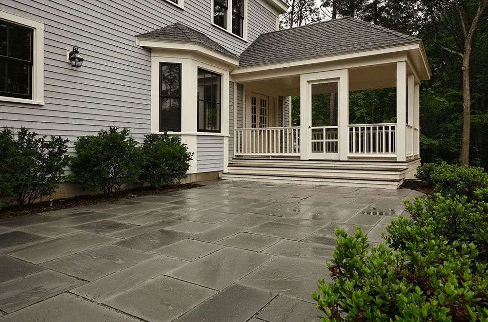 Gray patio with adjacent screened porch attached to a light gray house, with bushes.