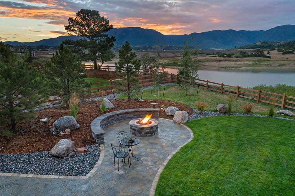 Fire pit with chairs on a patio overlooking a lake and mountains at sunset.