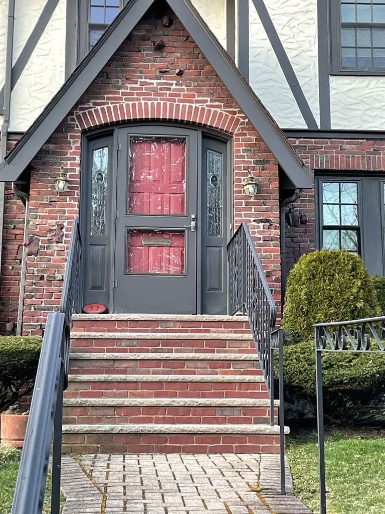 Red brick Tudor-style home entrance with steps leading up to a door, flanked by windows and shrubs.