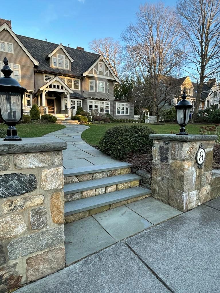 Stone walkway leading to a large grey house with a landscaped front yard under a blue sky.
