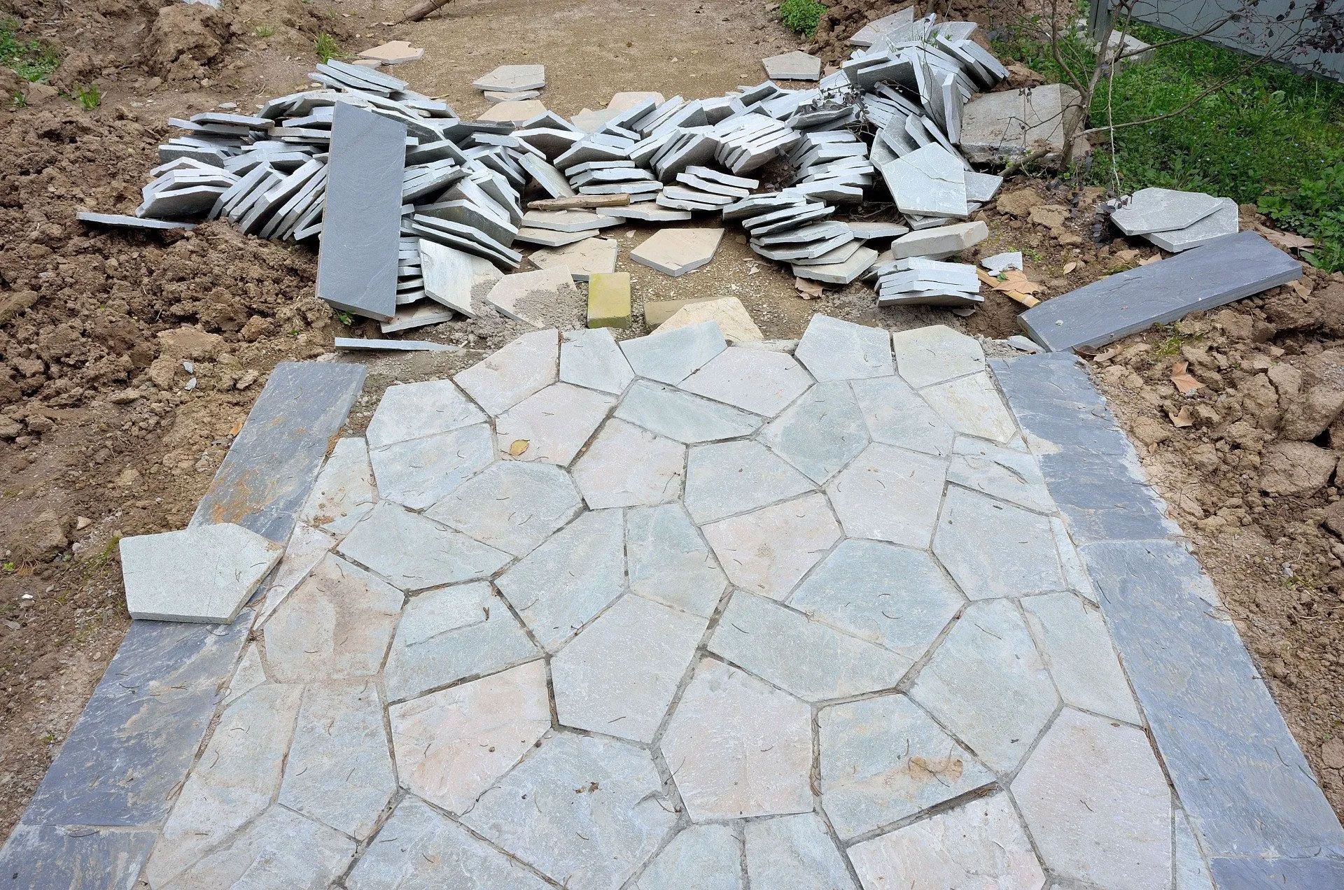 Pathway under construction, gray flagstone pavers laid in a random pattern, surrounded by dark border stones and loose stones.