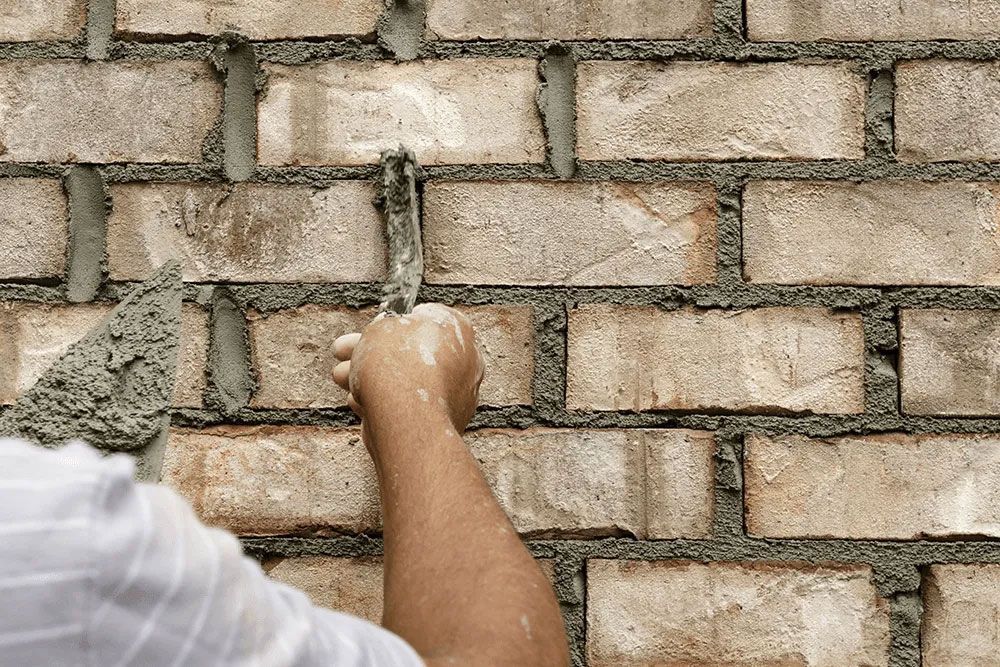 Person using trowel to apply mortar to brick wall.