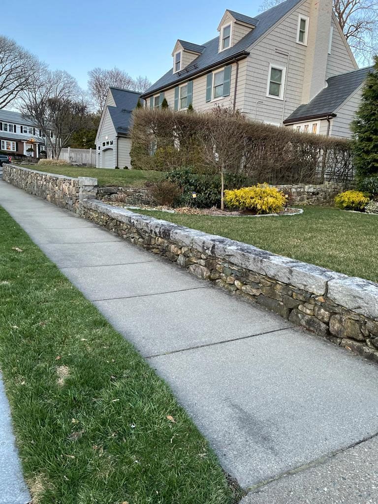 Sidewalk next to a stone wall and house with a gray roof. Green grass in foreground.