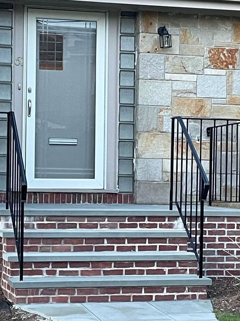 Brick steps lead to a white door with glass. Black handrails on both sides. Stone facade.