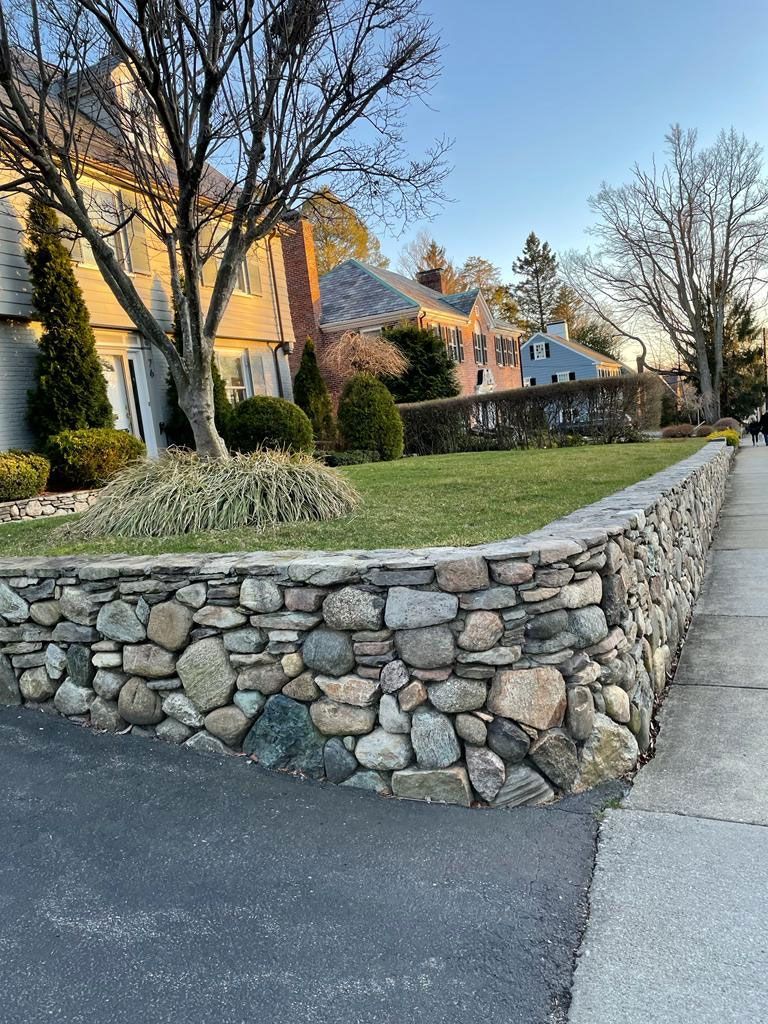 Stone wall borders a lawn in front of houses on a street with a paved sidewalk.