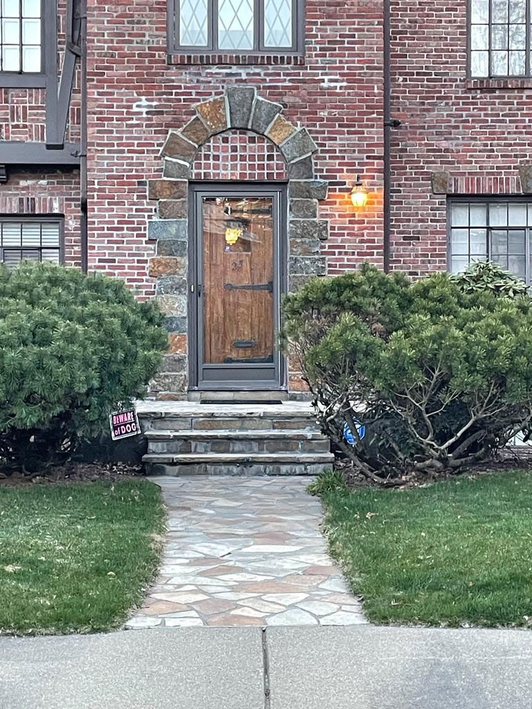 Brick home with stone archway over wooden door, approached by stone steps and path. Green bushes and grass.