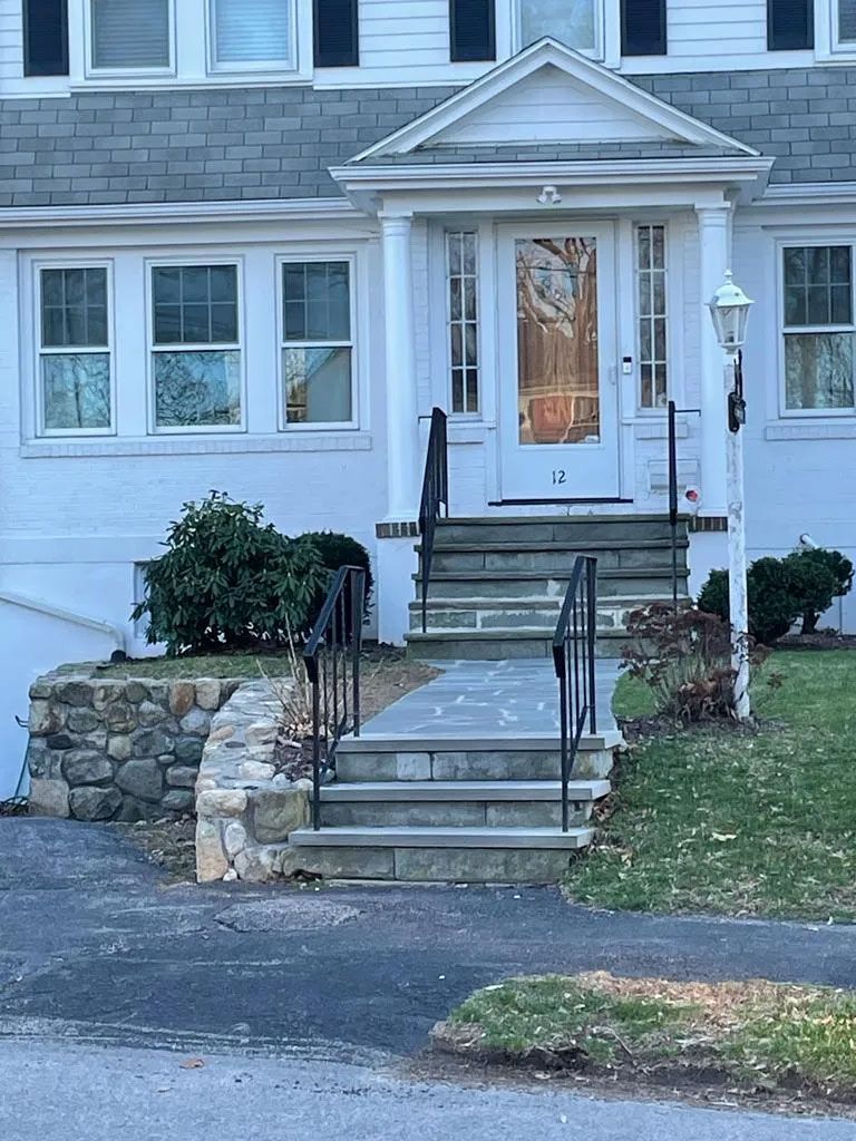White house with steps, stone wall, front door. Black railings. Blue-gray stone steps. Green grass.