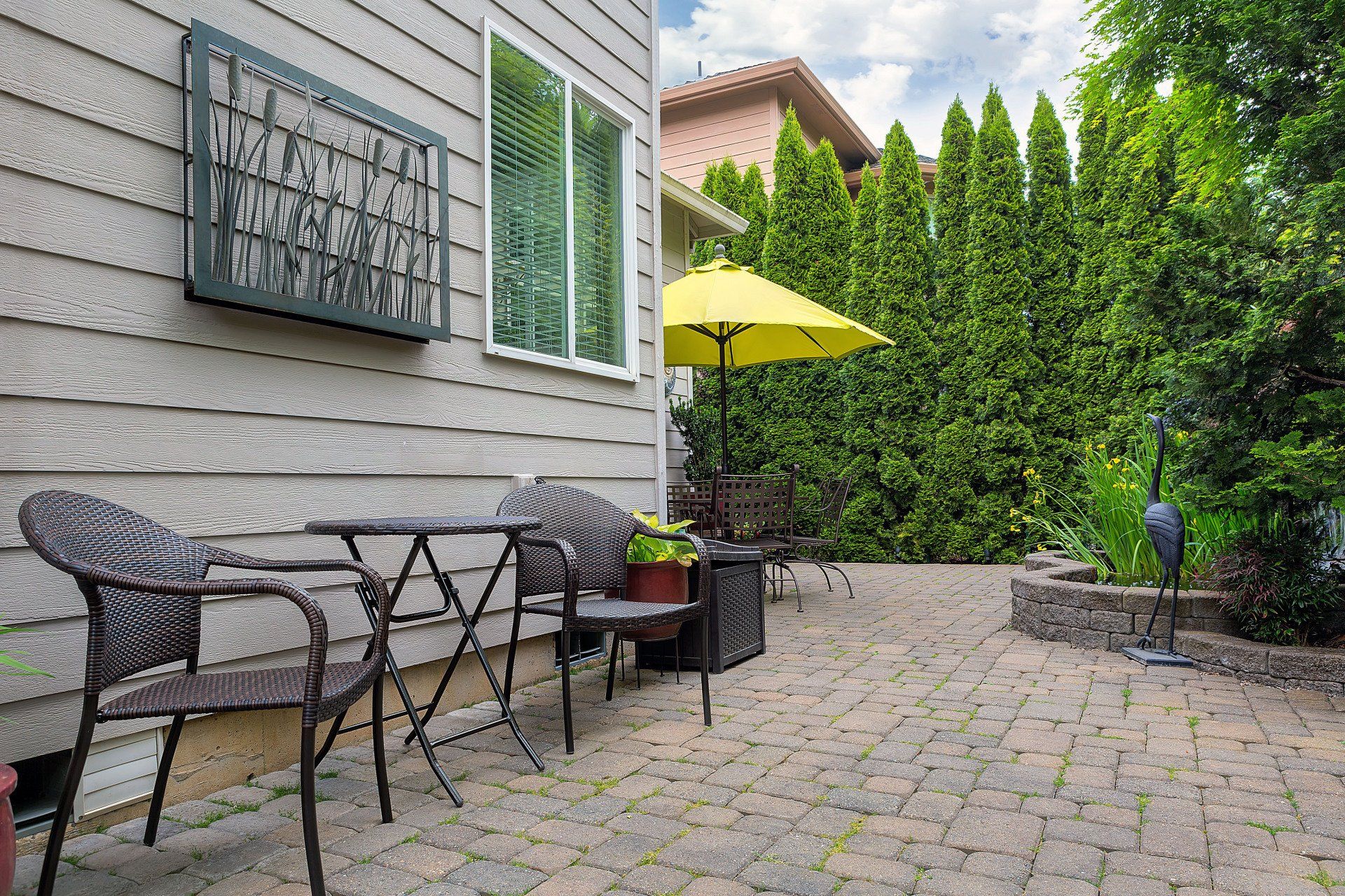 Patio with a small table, two chairs, and a yellow umbrella near a tall hedge.