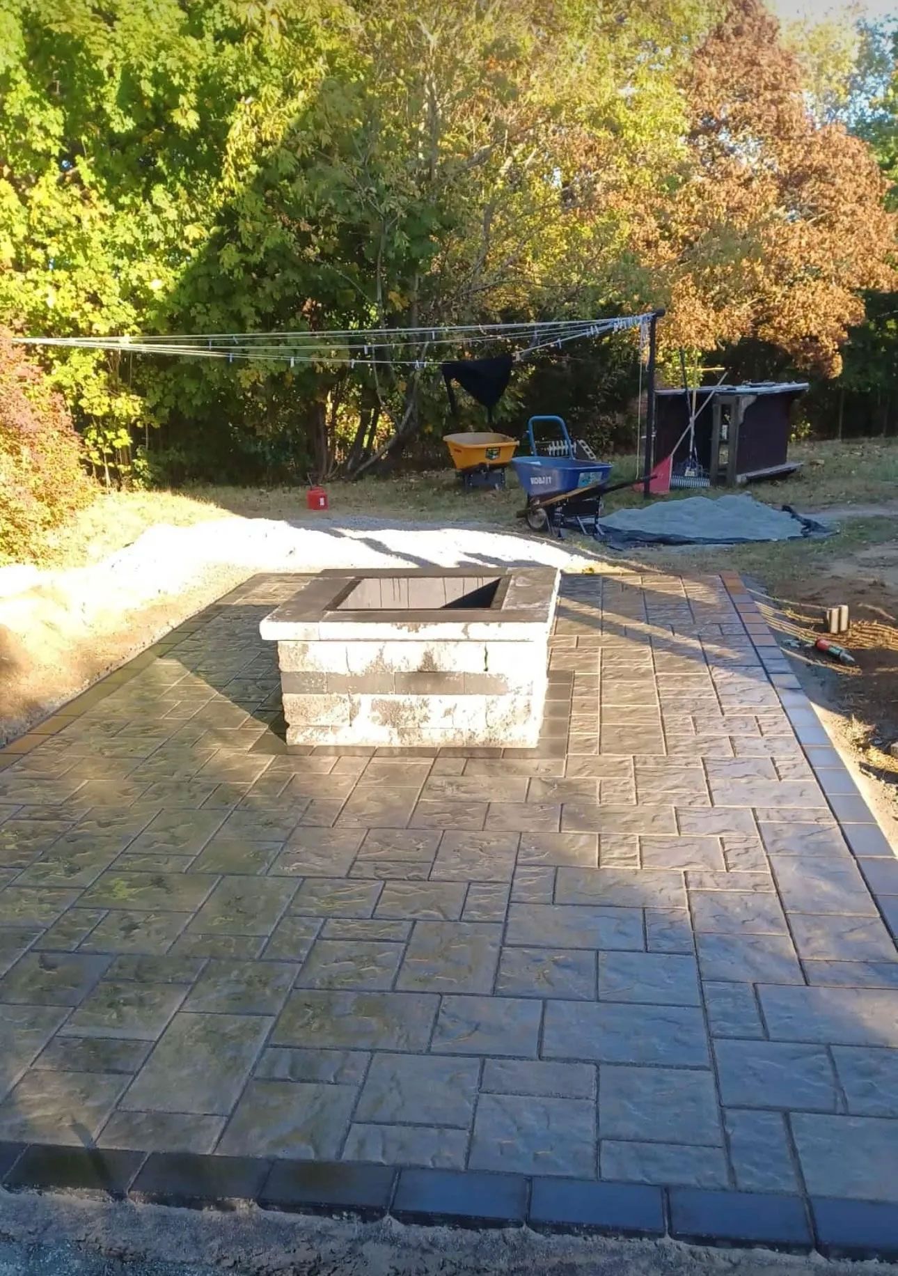 Concrete patio with fire pit, tools, and trees in background on a sunny day.