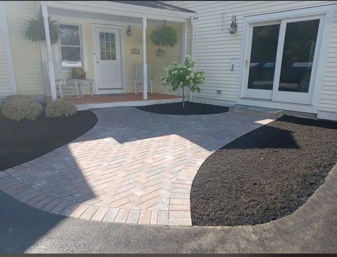 Brick pathway leads to a yellow house with a white door and sliding glass doors, surrounded by dark mulch.