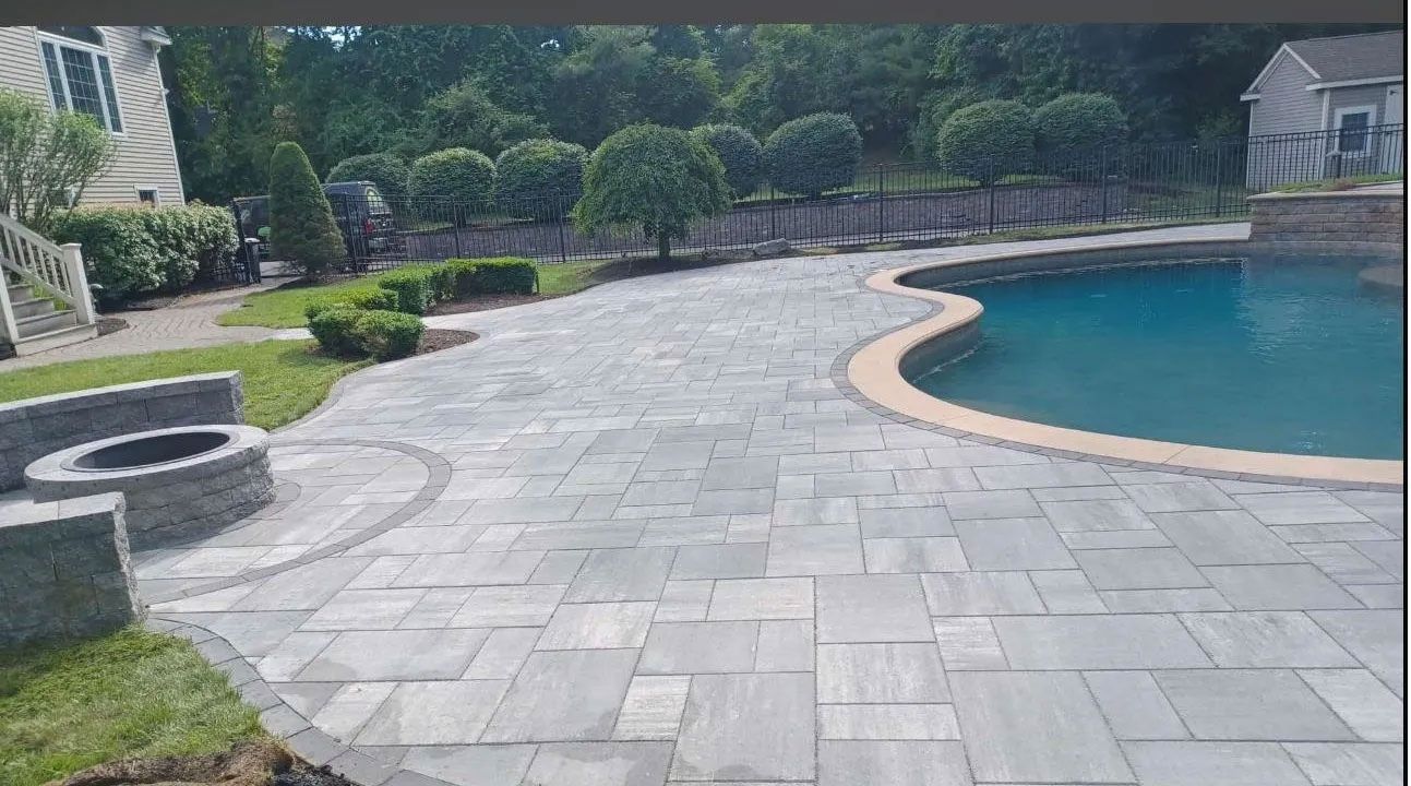 Stone patio surrounds a pool, with a fire pit, grass, and manicured bushes in the background.