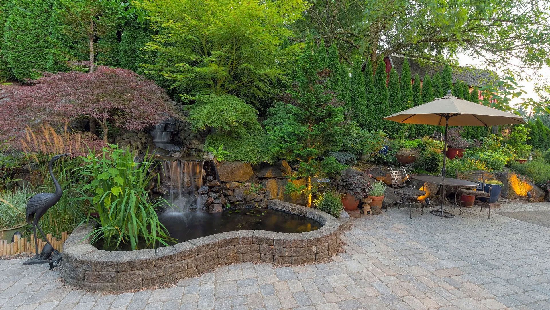 A serene backyard with a waterfall, pond, and patio seating under an umbrella. Lush greenery surrounds the scene.
