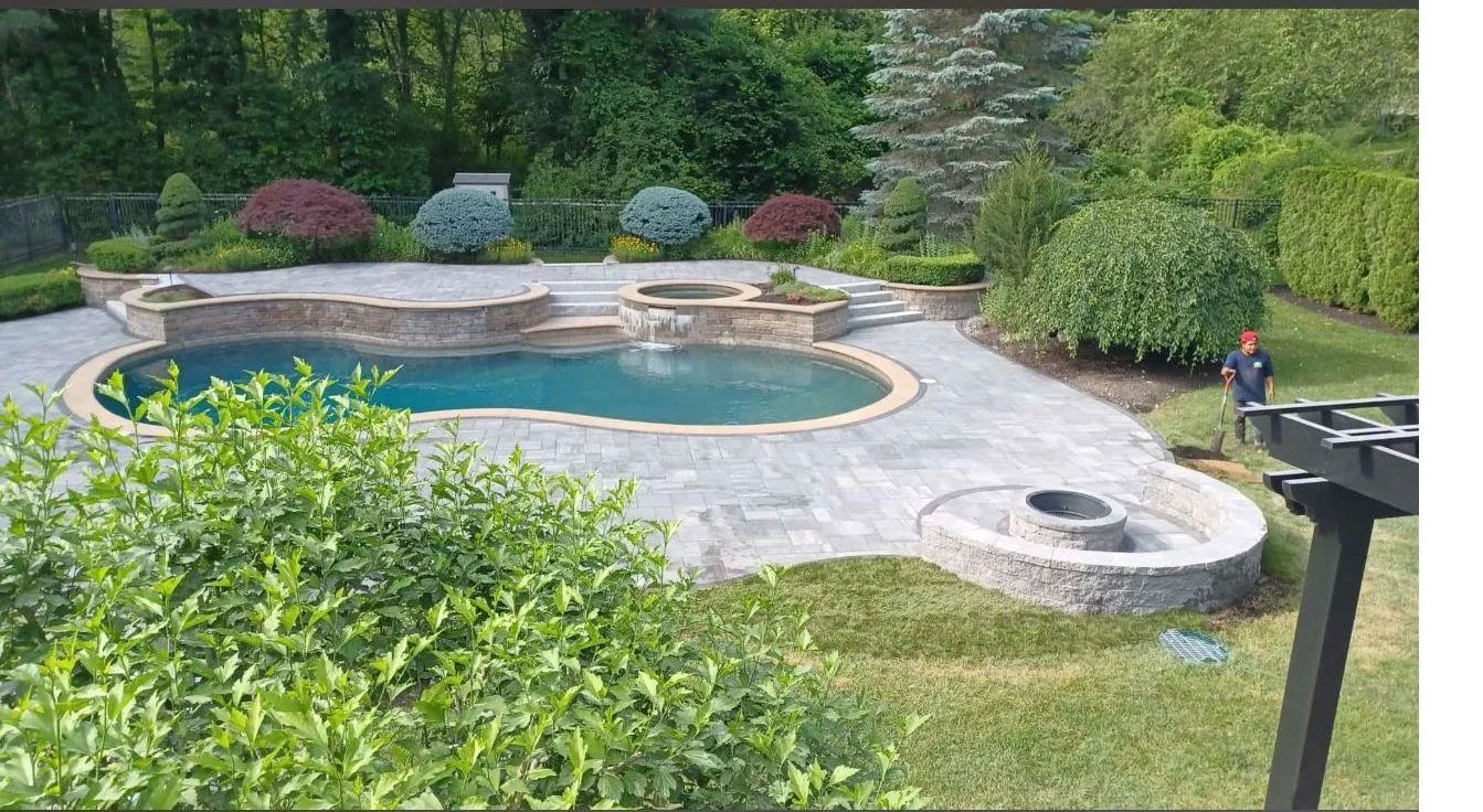 Pool and patio area in a backyard setting with greenery, trees, and a person.