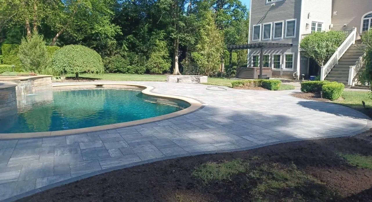 A backyard with a swimming pool, patio, and two-story house.  Green grass and trees surround the paved area.