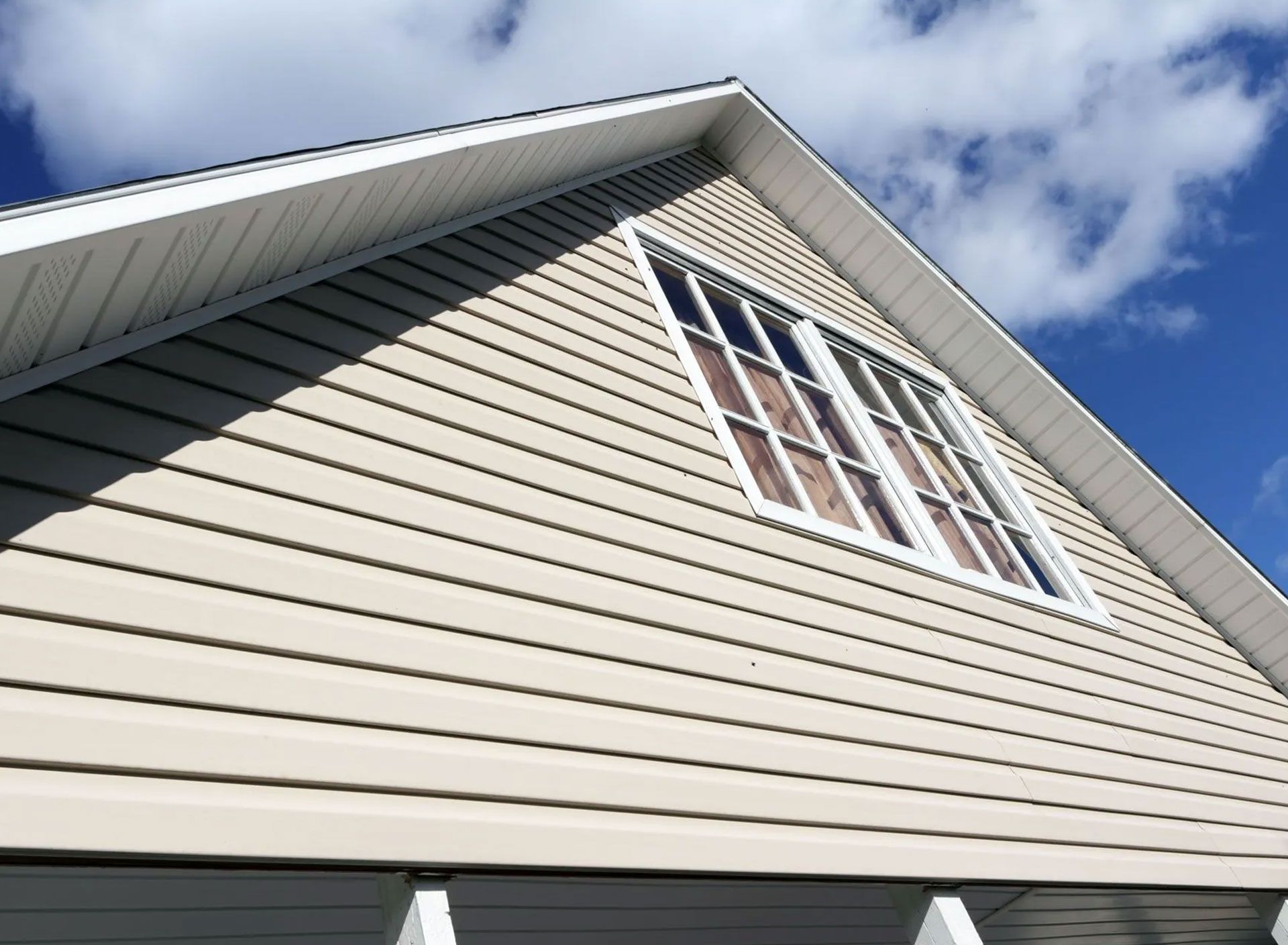 Beige siding on the angled side of a house with a window; a blue sky with clouds.