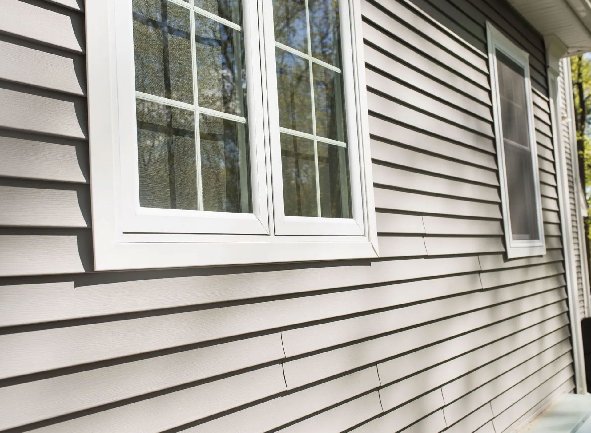 Gray siding on a house with white-framed windows. The sun is shining, visible tree tops in the background.