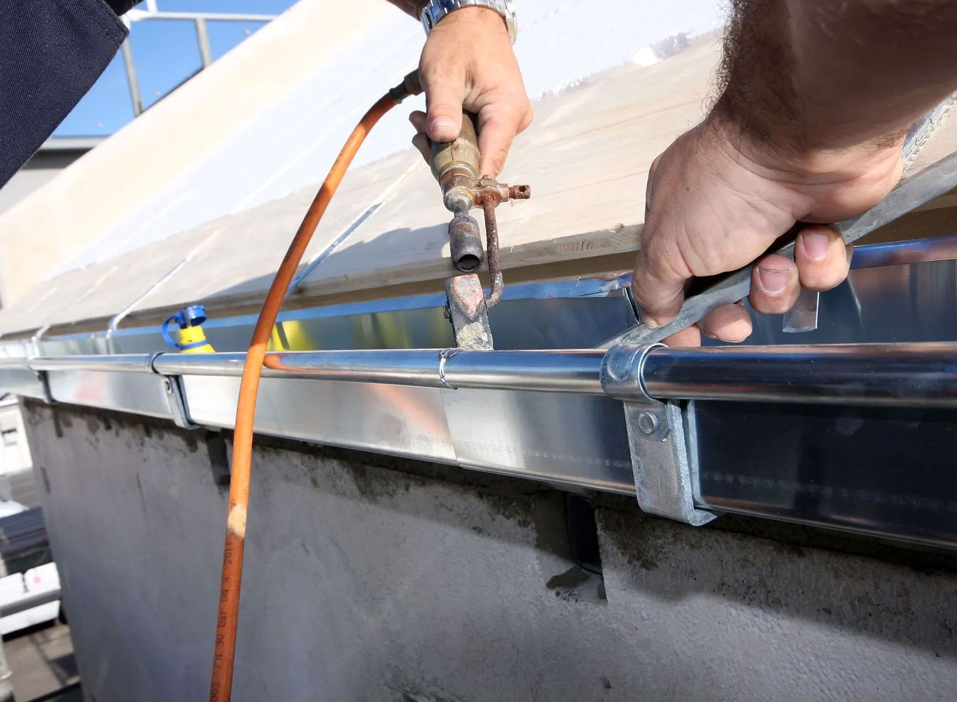 A person using a wrench to connect a hose to an outdoor faucet near a building's gutter.