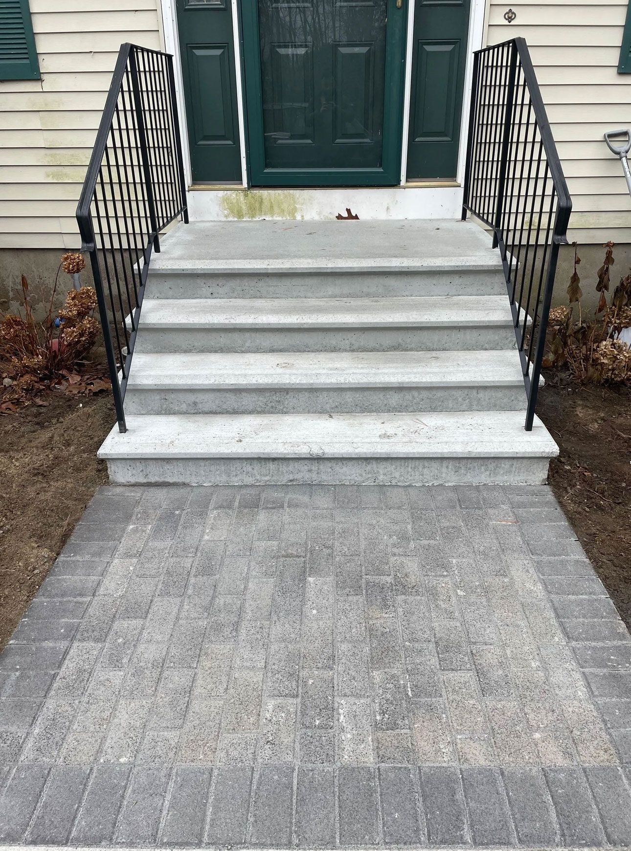 Grey stone steps lead to a green door with black iron railings, a brick walkway.