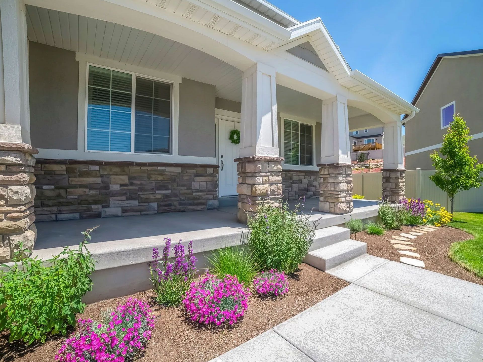 Beige house with a stone and white porch, purple flowers, and a walkway.