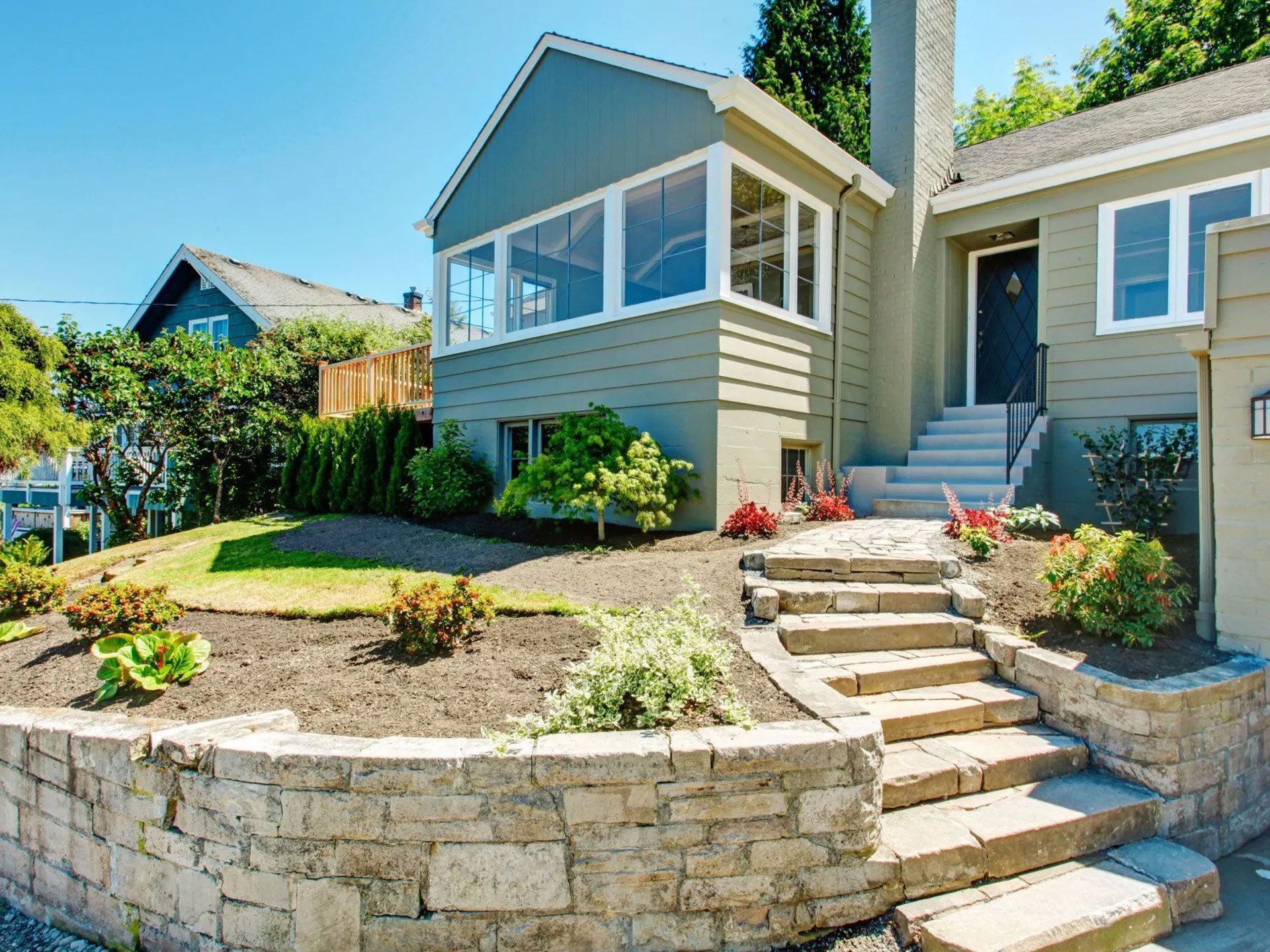 House with stone steps and retaining wall; blue-green siding; sunny day.