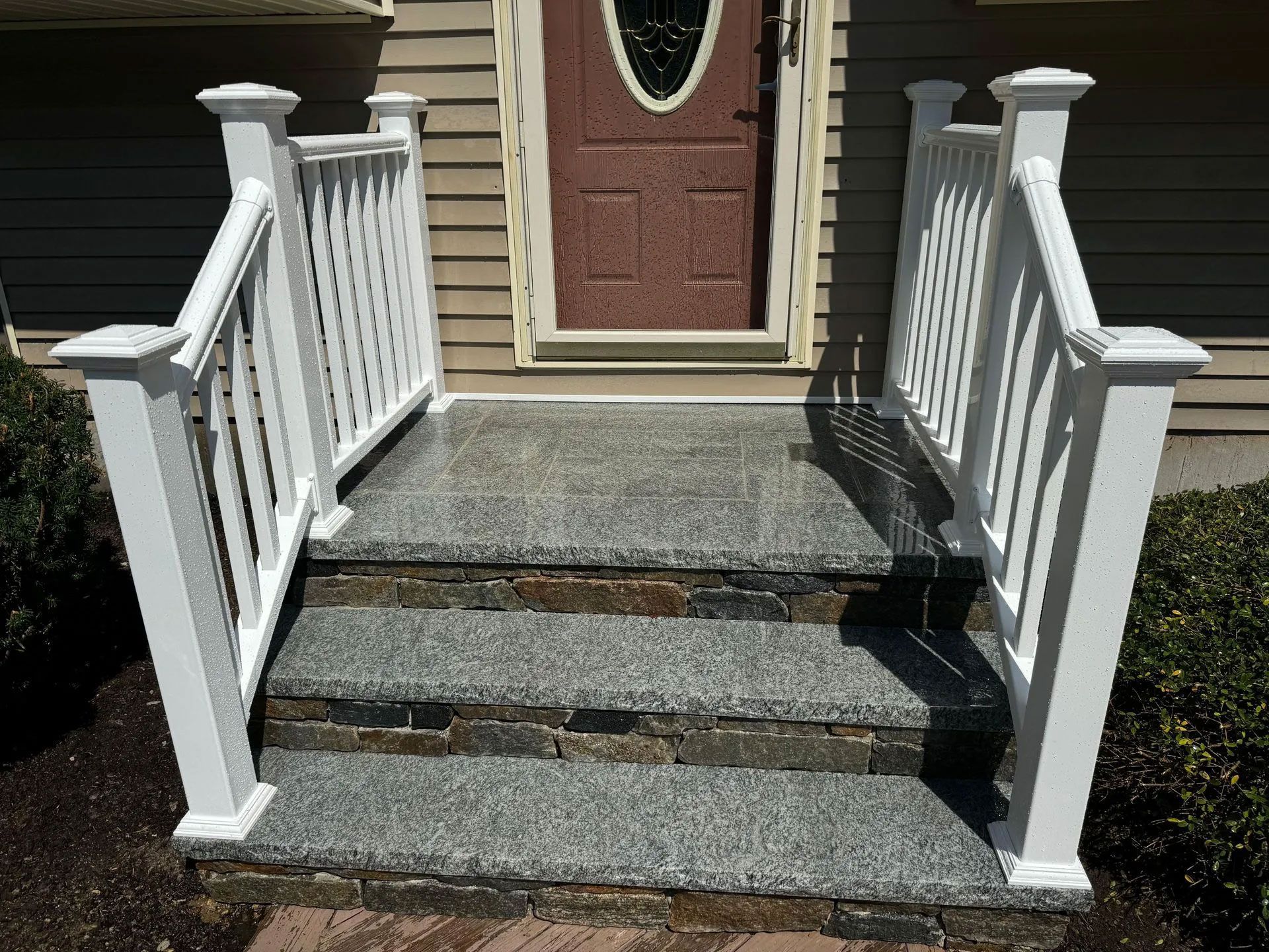 White-railed steps lead to a brown door. The steps are stone, the building has tan siding, and the sky is blue.