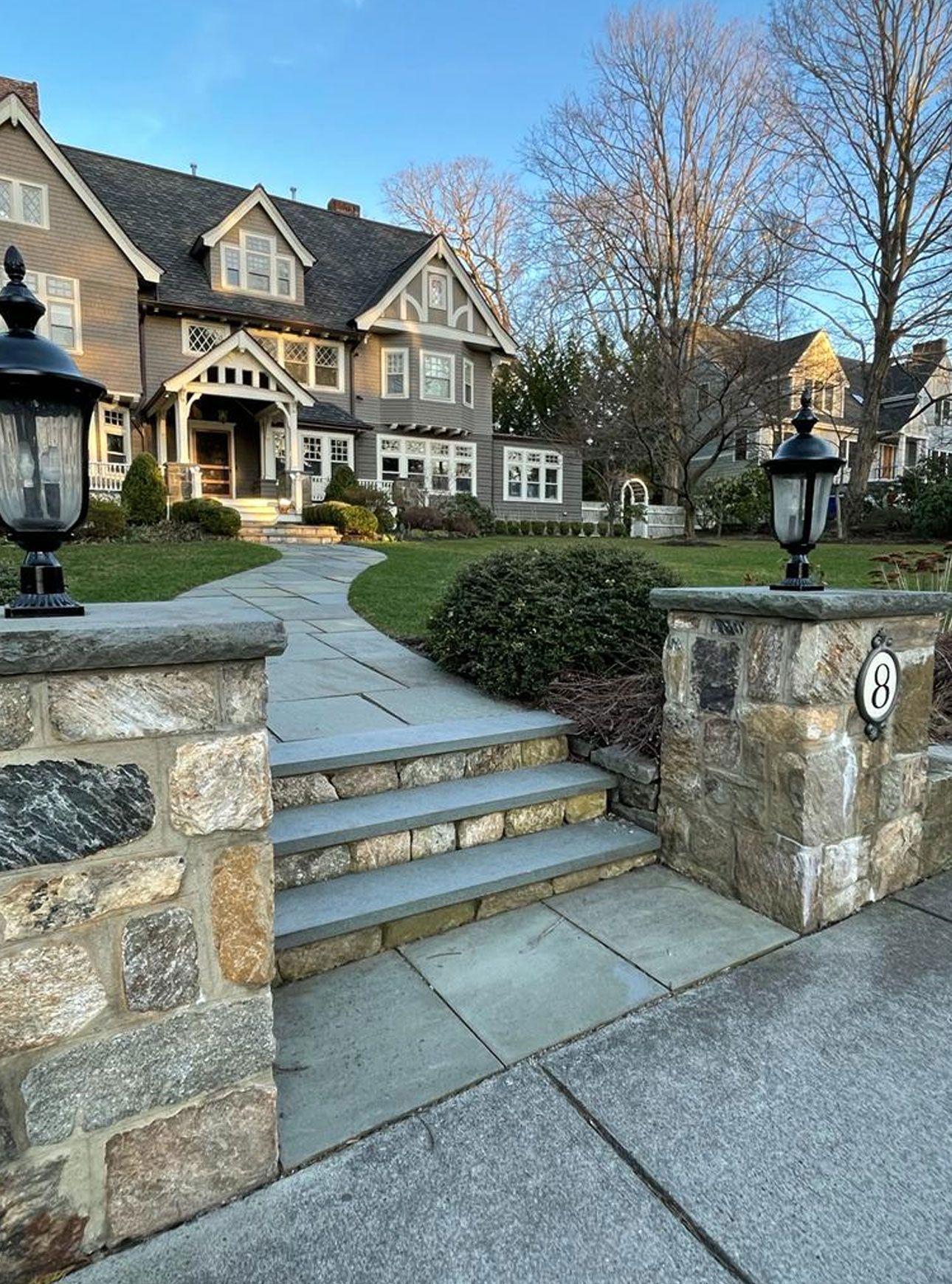 Stone walkway leading to a large gray house with manicured lawn, trees, and blue sky.