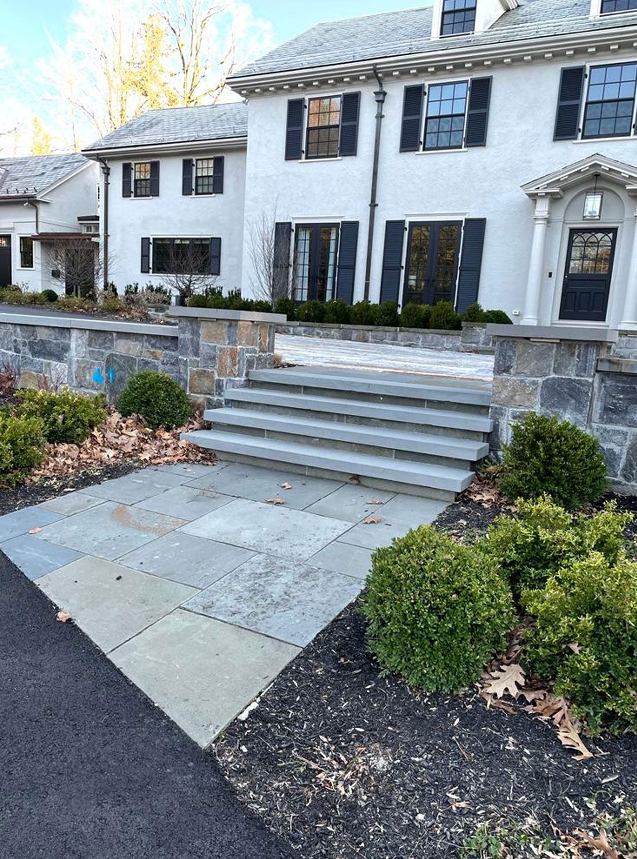 Stone steps lead to a white house with black shutters. Green bushes and stone walls surround the entrance.