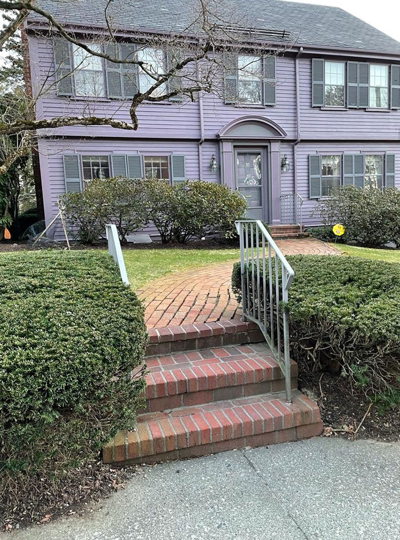 A lavender two-story house with brick steps, bushes, and a walkway.