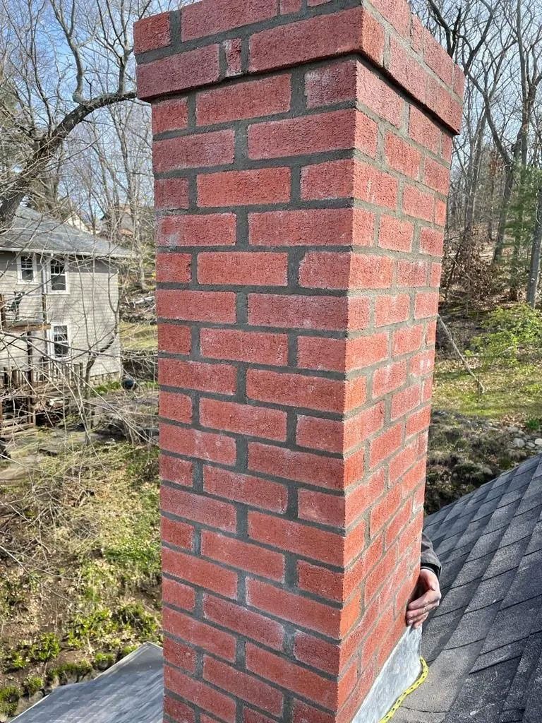 Brick chimney extending from a dark roof against a background of trees and a house.