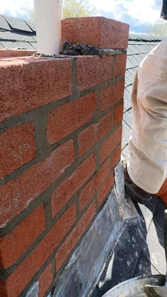 Bricklayer working on a red brick chimney with gray mortar. Roof in the background.