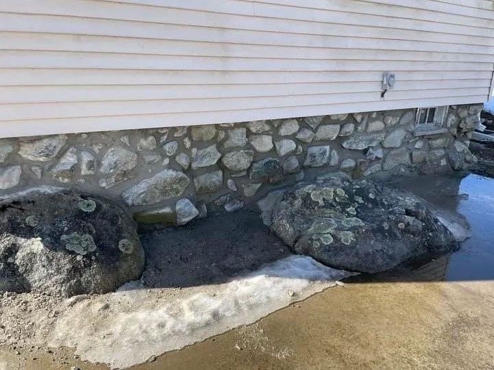 Base of a light-colored house with stone foundation and two large rocks, partially covered in snow.
