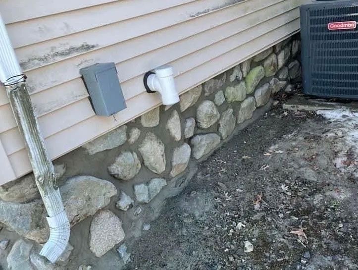 Stone foundation under beige siding with a white drainpipe and an HVAC unit.