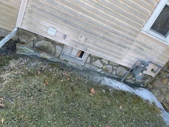 Stone foundation and light-colored siding on a building with a window and a small vent, beside grass.