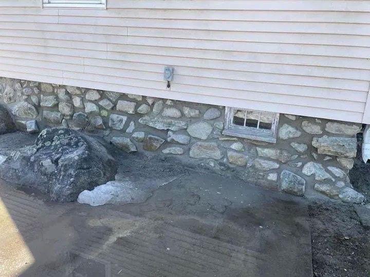 Stone foundation on a building's exterior with light-colored siding above. A rock pile and debris are visible in the foreground.