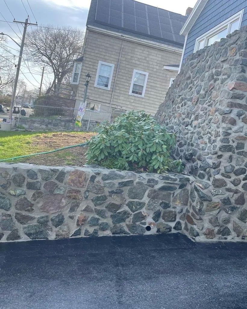 Stone wall beside paved area, with house and greenery in background.
