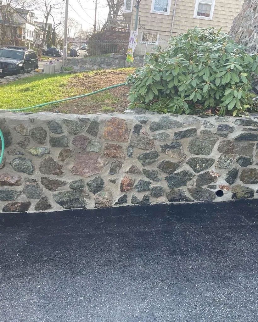 Stone retaining wall with green bush, asphalt driveway, and grassy area with parked cars.