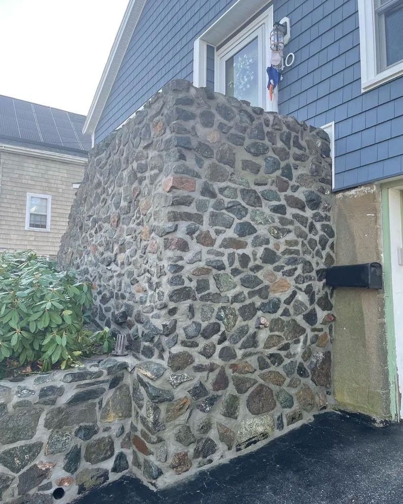 Stone wall supporting a blue house, with a green door and black mailbox, and a brick building to the left.