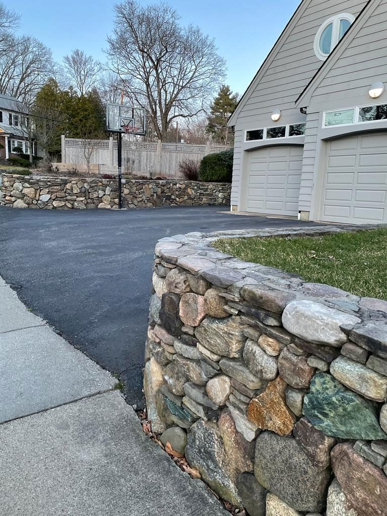 Stone retaining walls border a black asphalt driveway leading to a two-car garage; a basketball hoop sits in the distance.