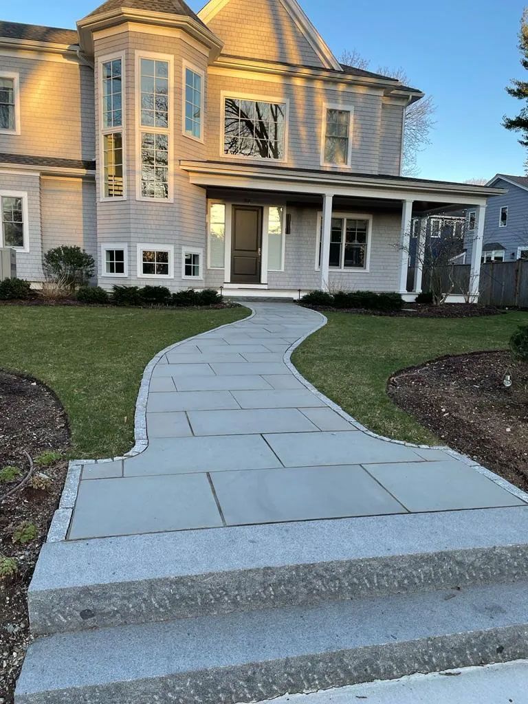 Stone path leading to a light grey house with a covered porch and a rounded turret, green grass, and blue sky.