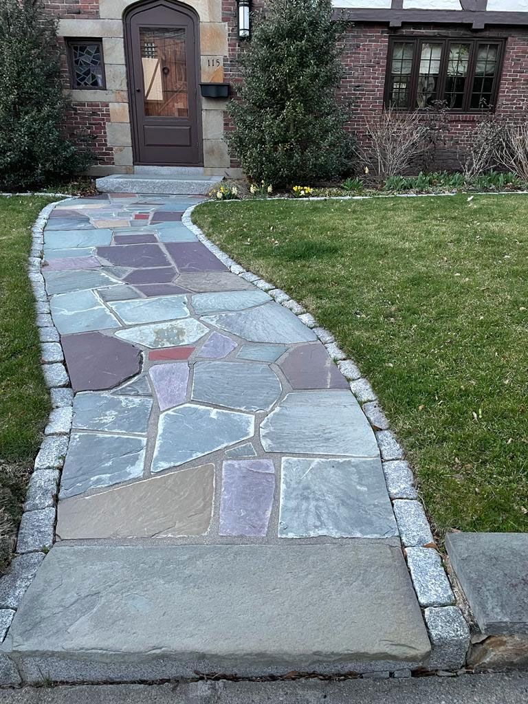 Stone walkway leading to a brown door, flanked by grass and shrubbery, with a brick house facade.