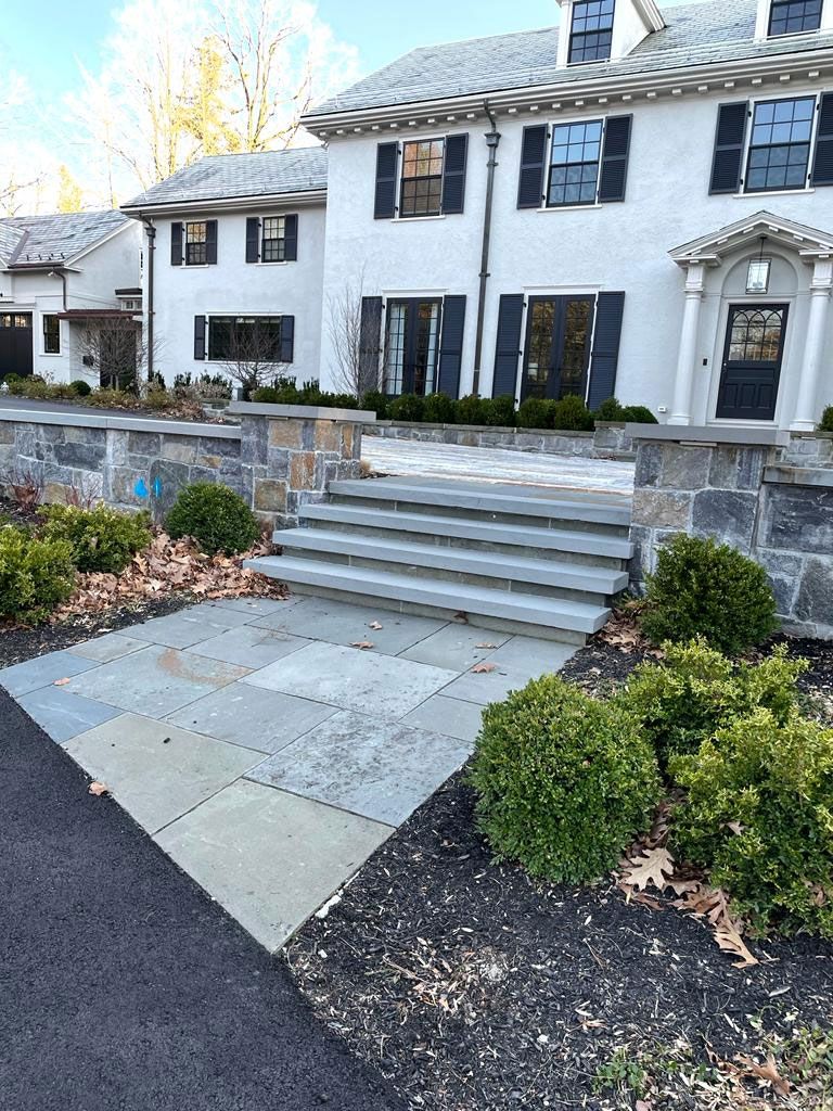 Stone steps leading up to a light-colored house with black shutters; stone retaining walls and greenery.