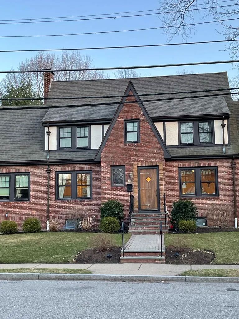 Brick two-story house with black trim, dark roof, front door, and small front yard with grass and bushes.