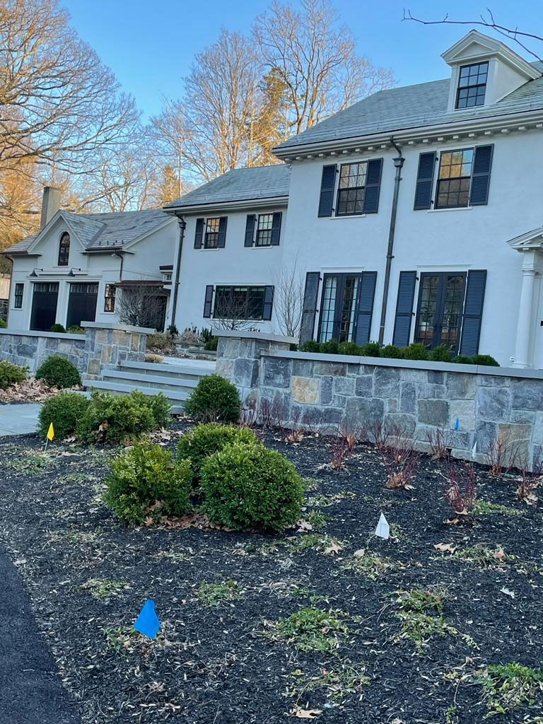 Large white house with black shutters and stone wall in front. Bushes and dark mulch in the foreground.
