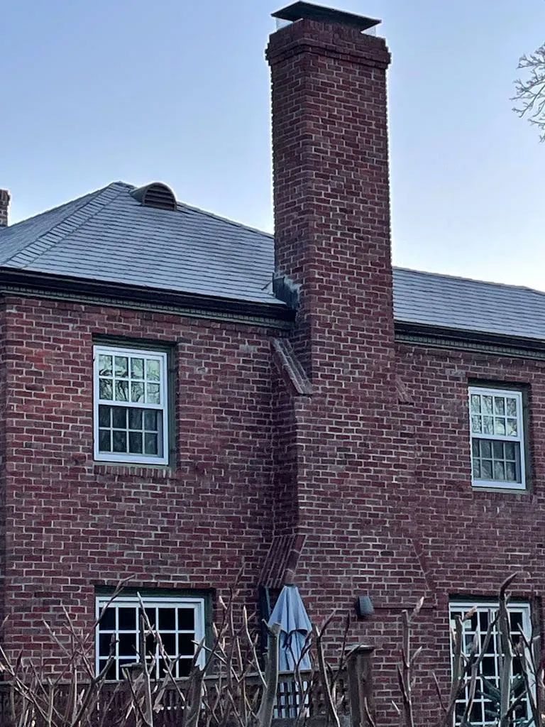 Brick house with chimney; white-framed windows; blue umbrella in yard; gray roof.