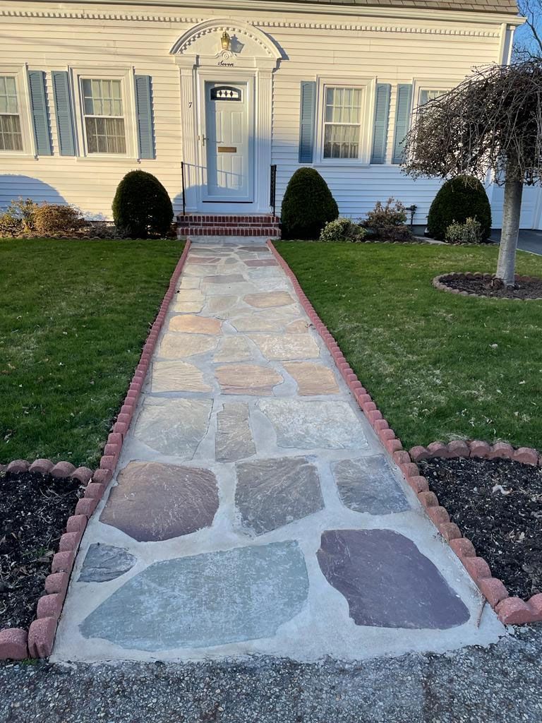Stone walkway leading to a white house with blue shutters, flanked by grass and flowerbeds.