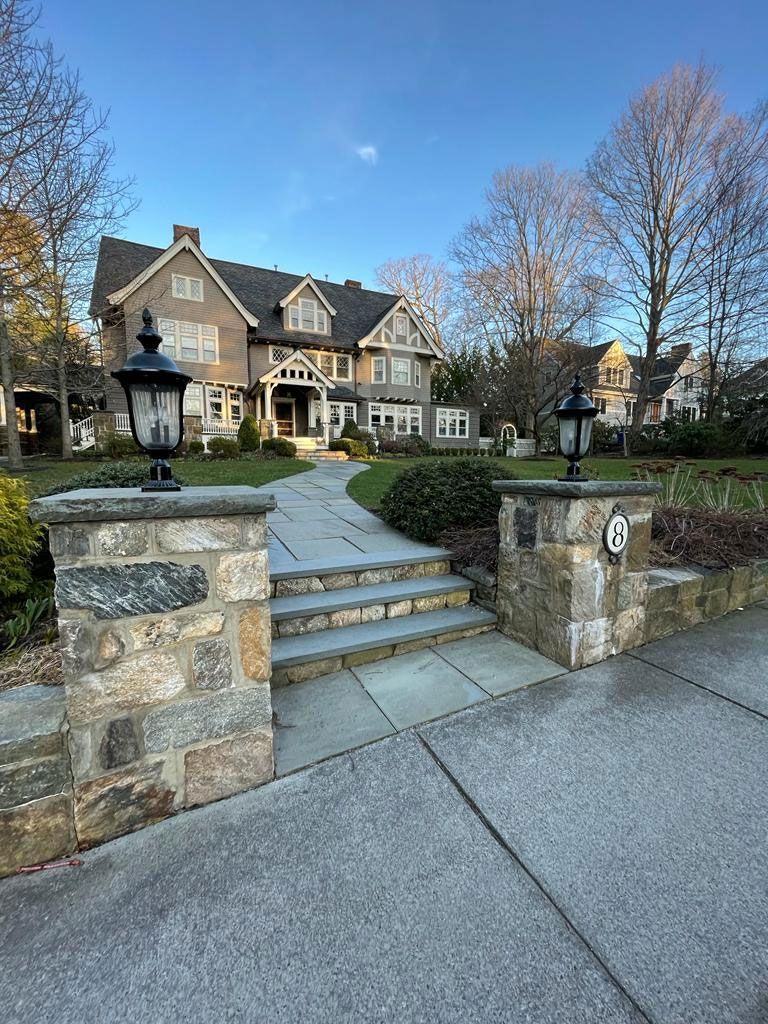 Stone pillars with lanterns flank a stone path leading to a large gray house with a green lawn under a blue sky.
