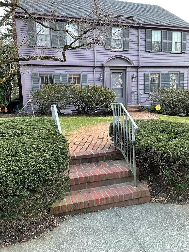 Purple house with gray shutters. Brick steps lead to the front door, flanked by green bushes.
