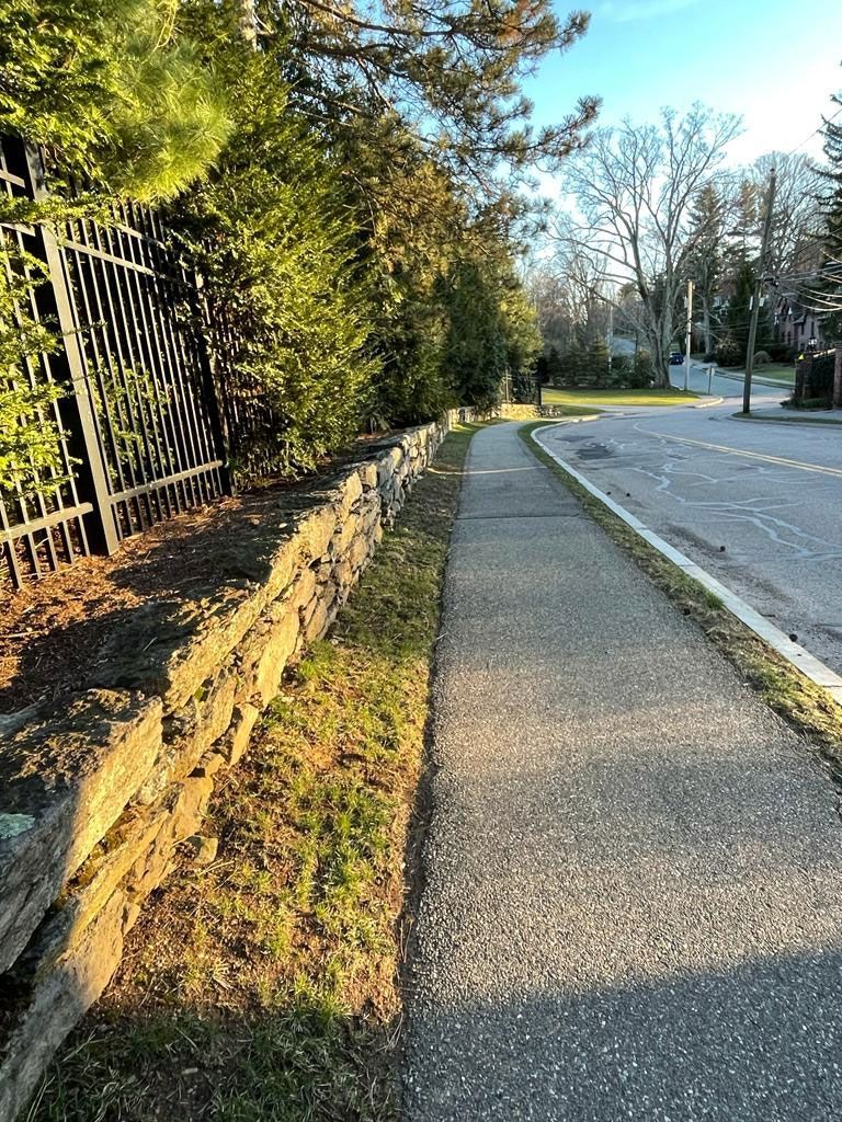 A sidewalk next to a wall and road, with trees and a fence on the left.