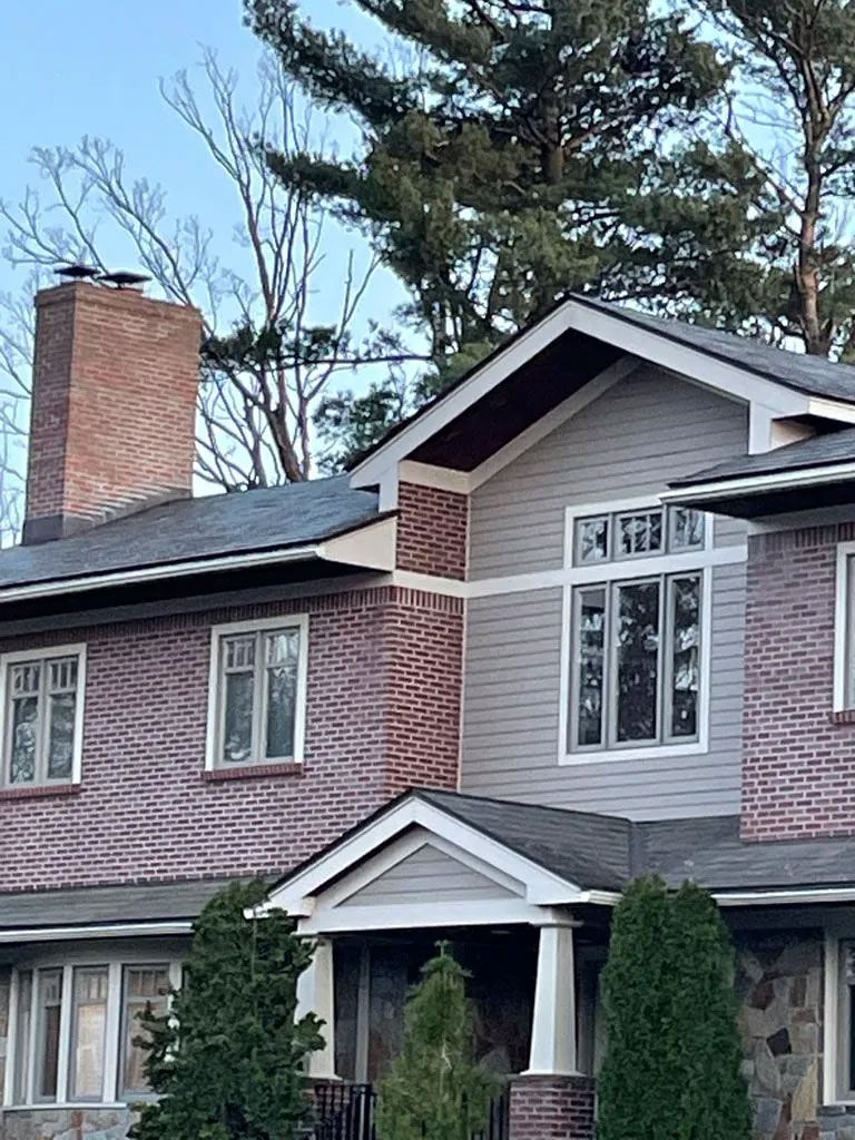 Two-story brick and gray house with a chimney and various windows, trees in the background.